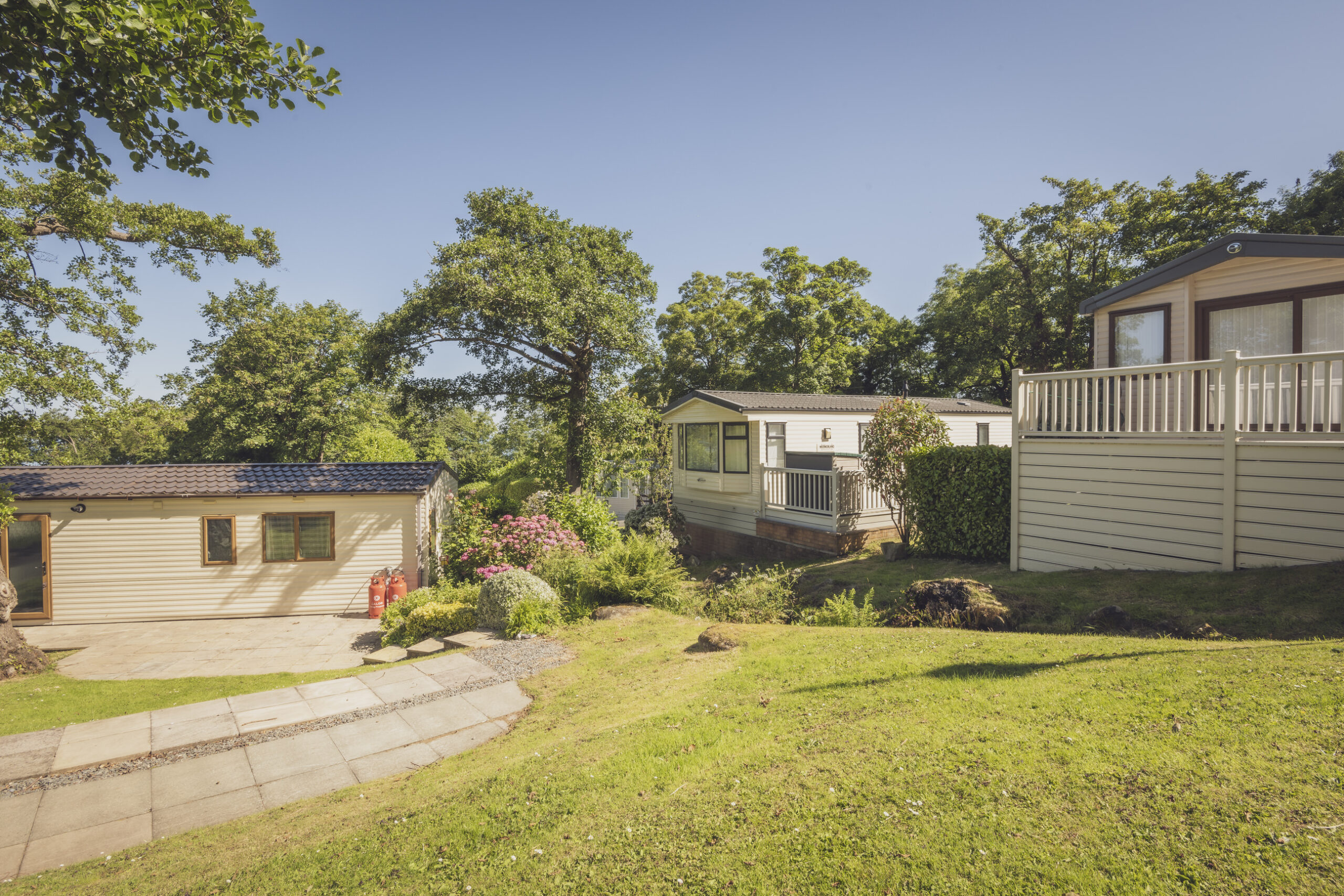 Static caravans on a grassy, tree-lined plot, with paved paths, a patio, gas canisters, and flowering shrubs in the foreground under a sunny sky.