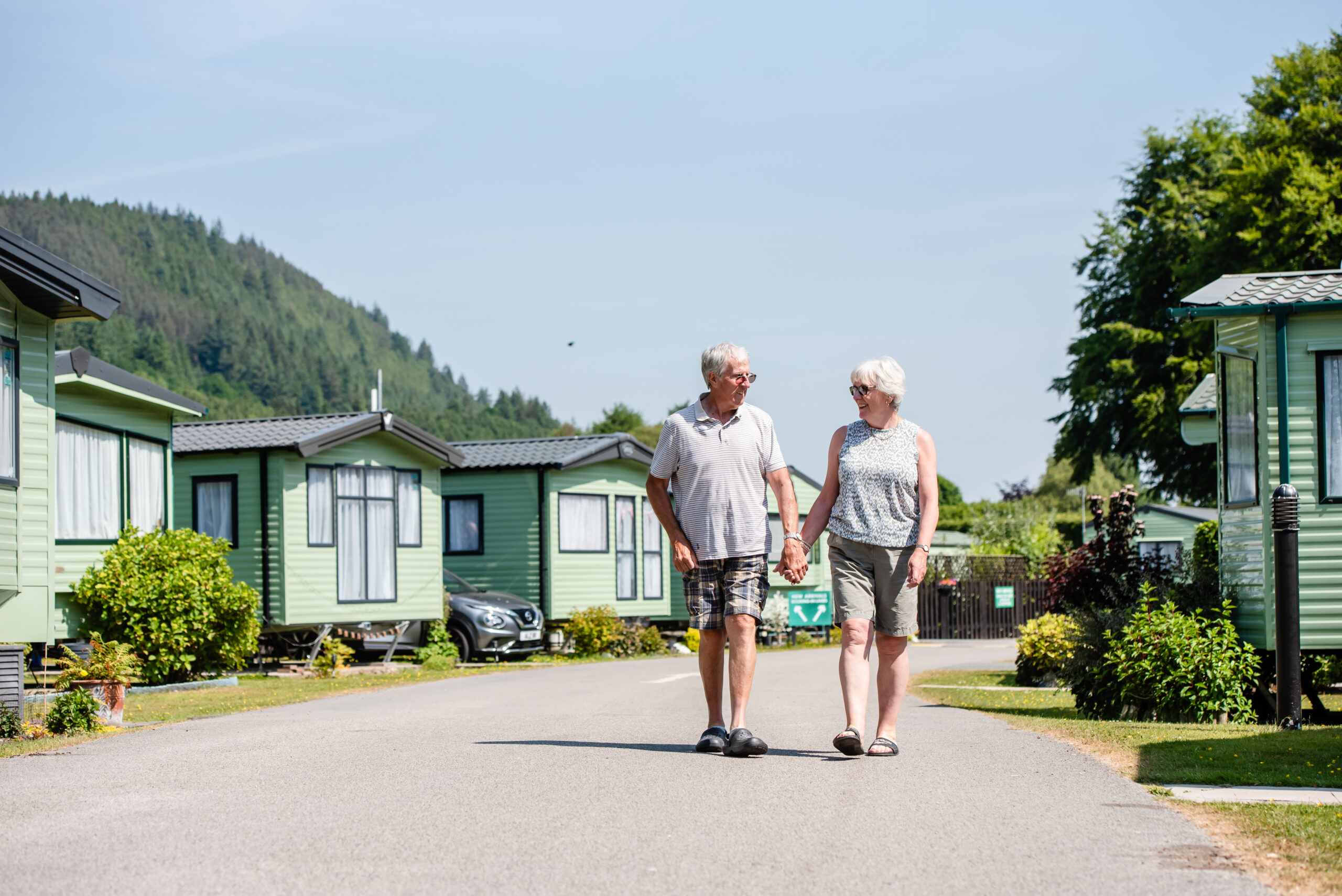 An older couple smile at each other while holding hands, walking down a paved lane between rows of green static caravans at a holiday park, with a tree-covered hill in the background.