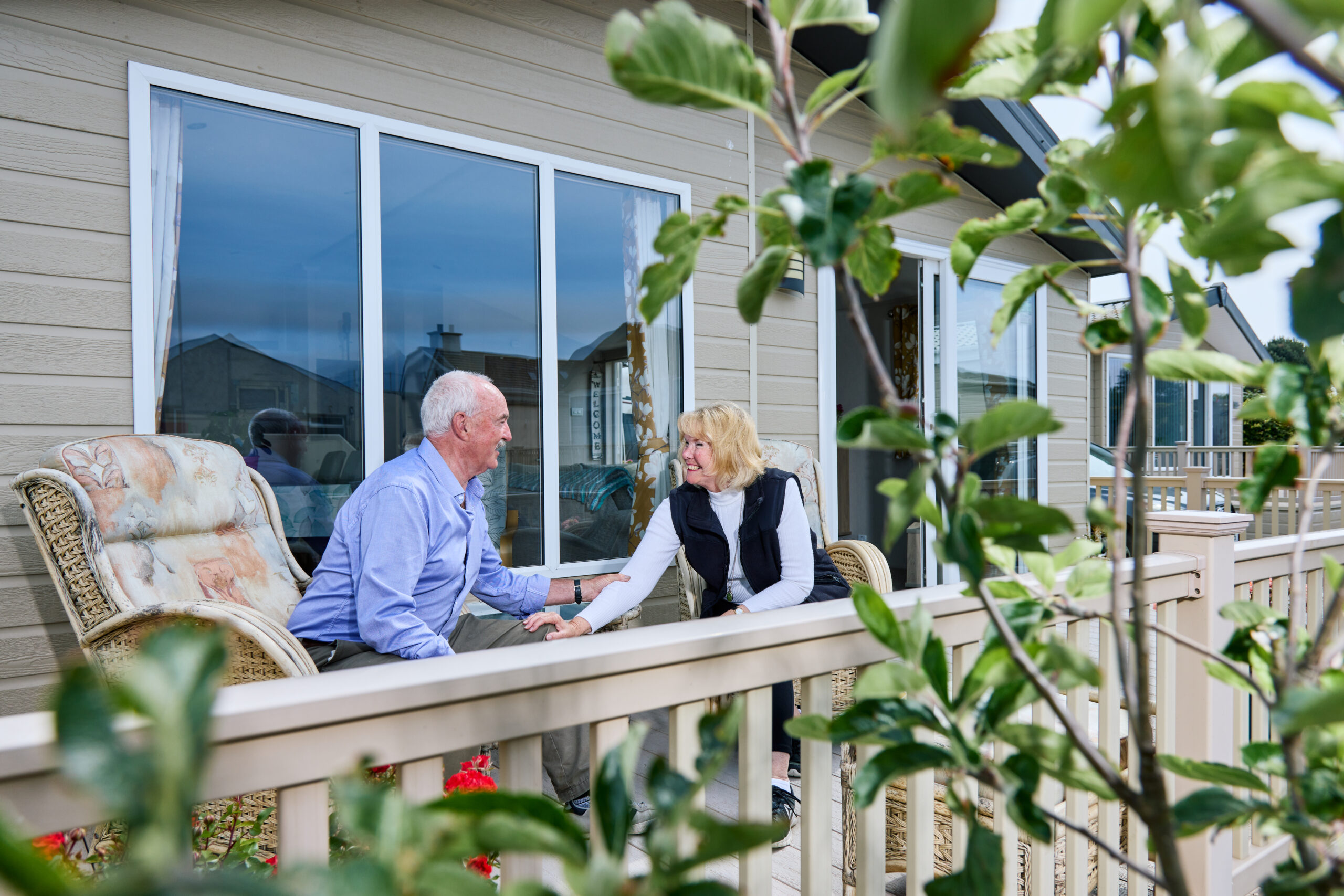 A couple sit facing each other on wicker chairs on the decked veranda of a Willerby lodge. The man wears a light blue shirt and the woman a white top and dark gilet. Green foliage and red flowers frame the foreground, with additional lodge accommodation visible in the background.