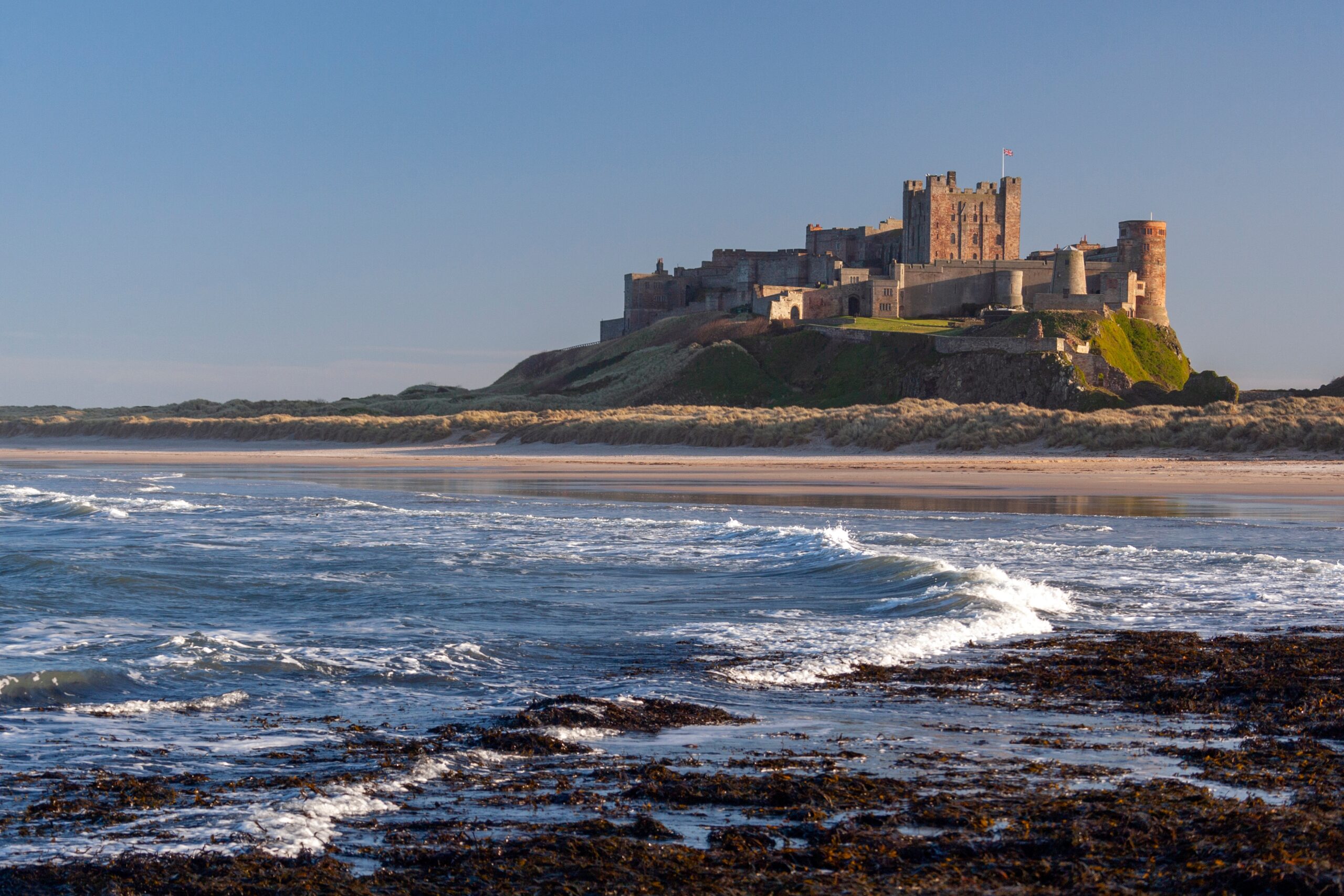 Bamburgh Castle rising dramatically above the Northumberland coastline, viewed from the beach below. The medieval stone fortress sits atop a rocky, grass-covered basalt outcrop, its keep and towers bathed in warm golden sunlight against a clear blue sky.