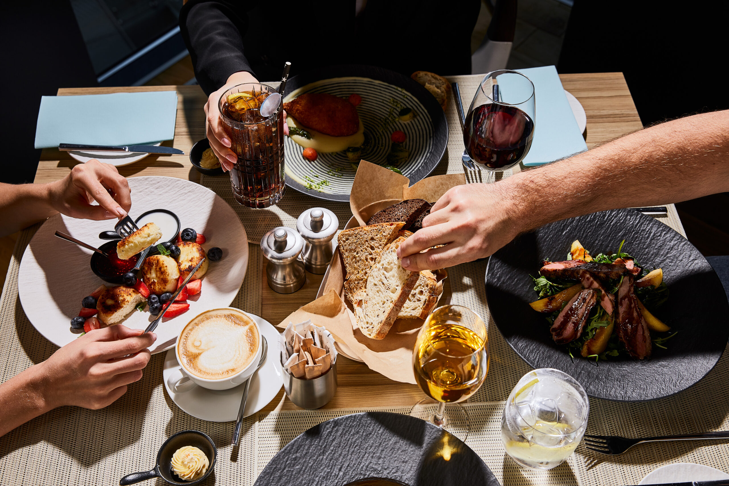 An overhead shot of people sharing a meal at a restaurant table, with a spread of dishes, bread and drinks. 