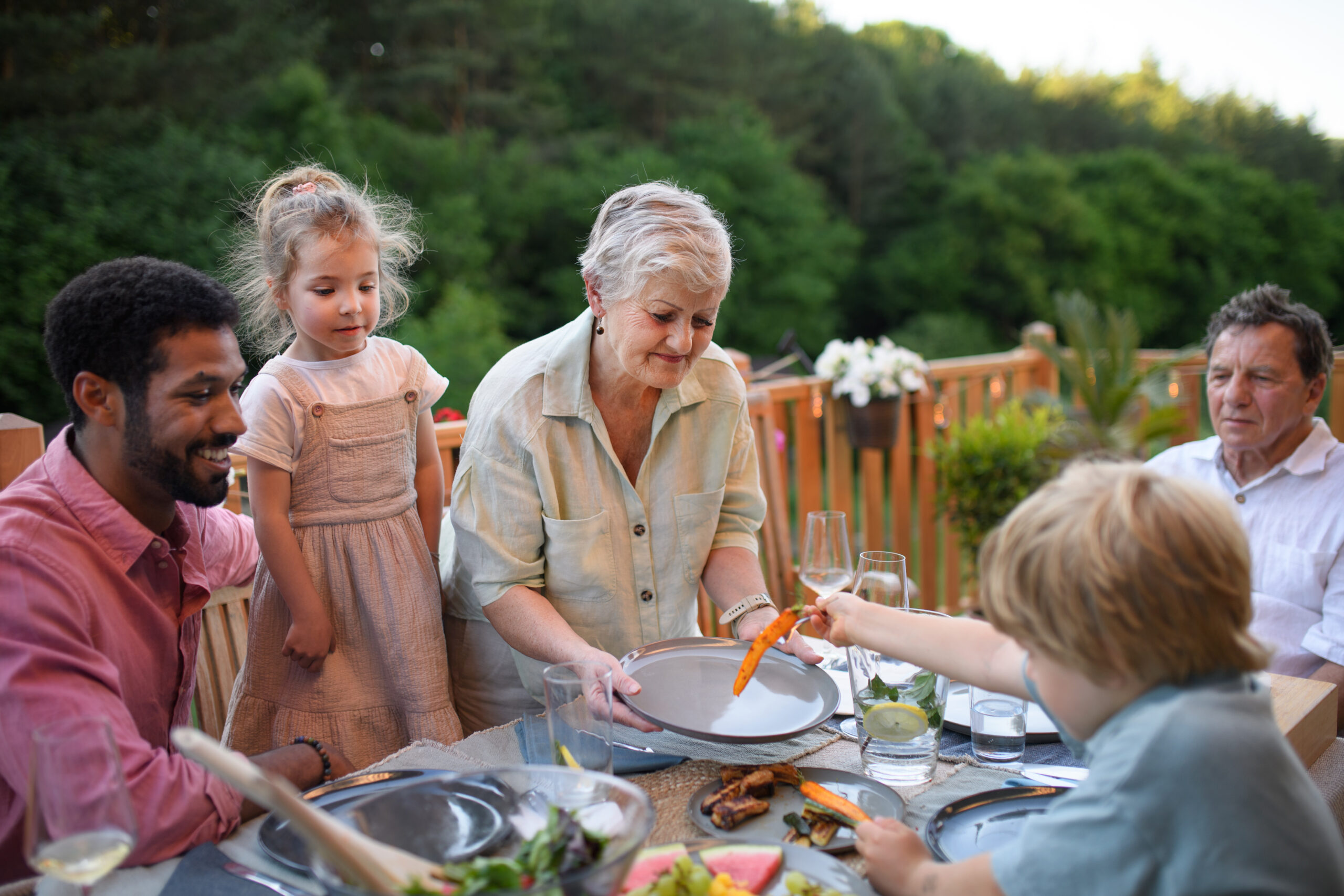 A multigenerational family gathered around an outdoor table, sharing a warm meal together on a wooden deck with greenery in the background.