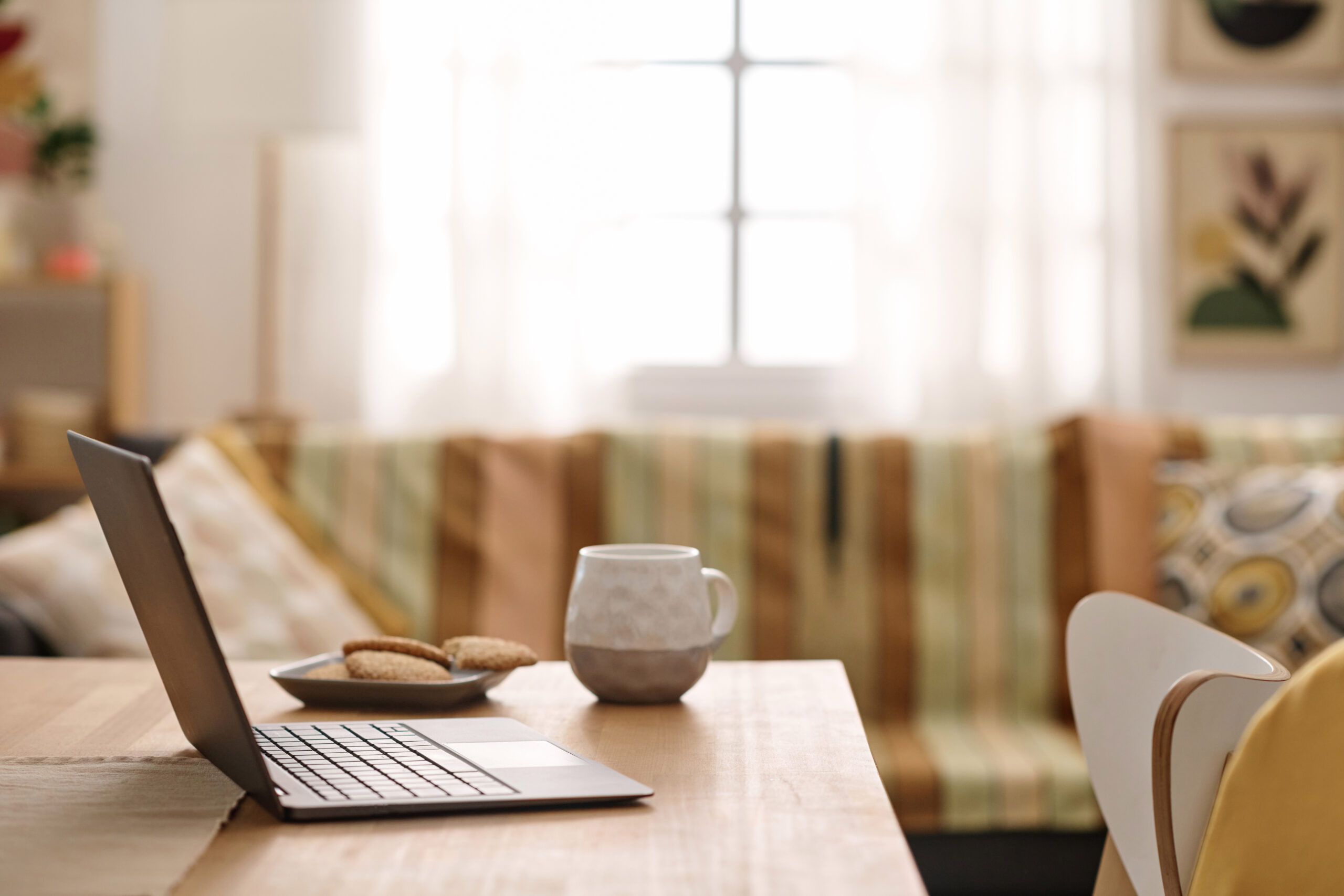 An open laptop on a wooden dining table at home, with a mug and a small plate of biscuits beside it, and a cosy living room in the background.