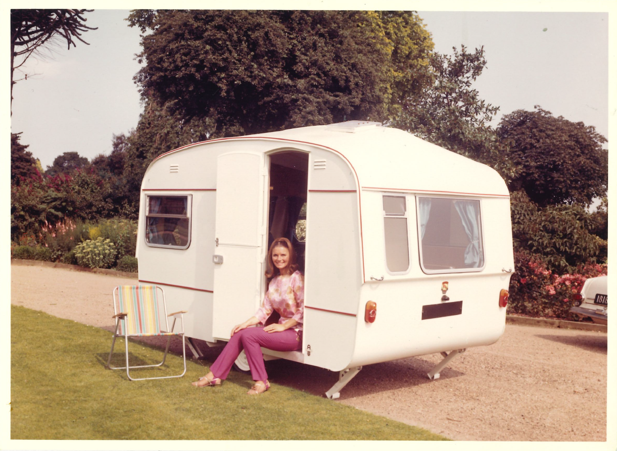 A vintage photograph from the 1960s of a woman sitting in the doorway of a white Willerby caravan, with a striped folding chair on the grass beside her.