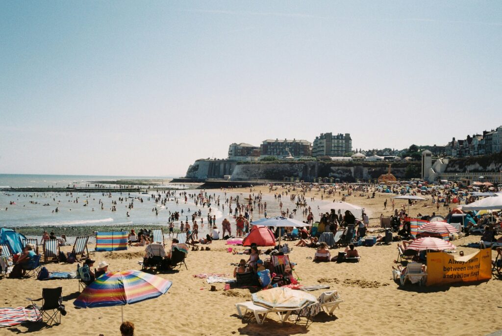 A packed British beach on a sunny day, with families relaxing in deckchairs and paddling in the sea beneath a clear blue sky.