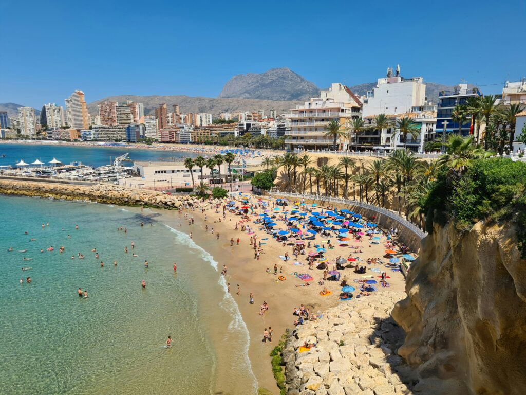 An aerial view of a busy Benidorm beach on a sunny day, with colourful umbrellas, turquoise water, palm trees and the resort's iconic skyline in the background