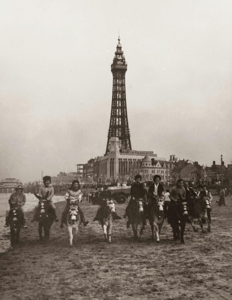 A vintage photograph of families enjoying donkey rides on a busy Blackpool beach, with Blackpool Tower dominating the skyline behind them