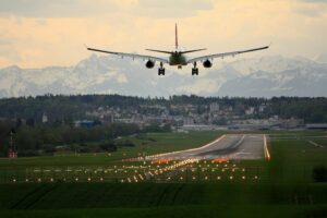 A passenger aircraft coming in to land at dusk, with snow-capped mountains visible in the distance and runway lights stretching ahead