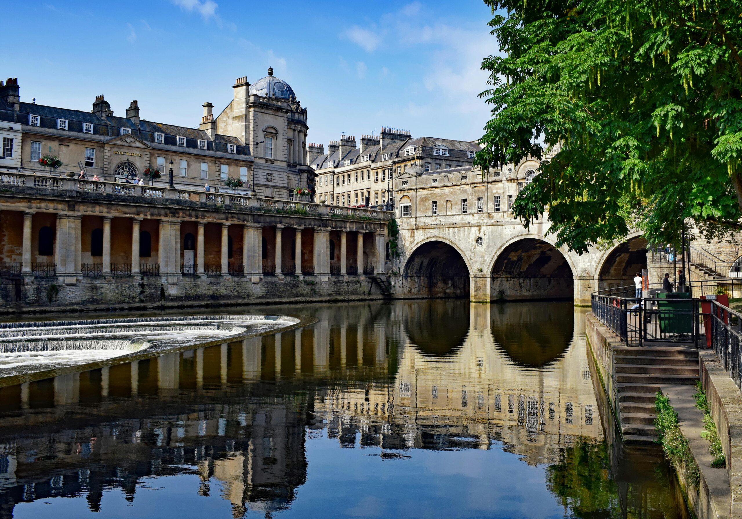 Pulteney Bridge and the River Avon in Bath, with the honey-coloured Georgian architecture reflected in the calm water below and a horseshoe weir curving across the river to the left, on a bright summer's day