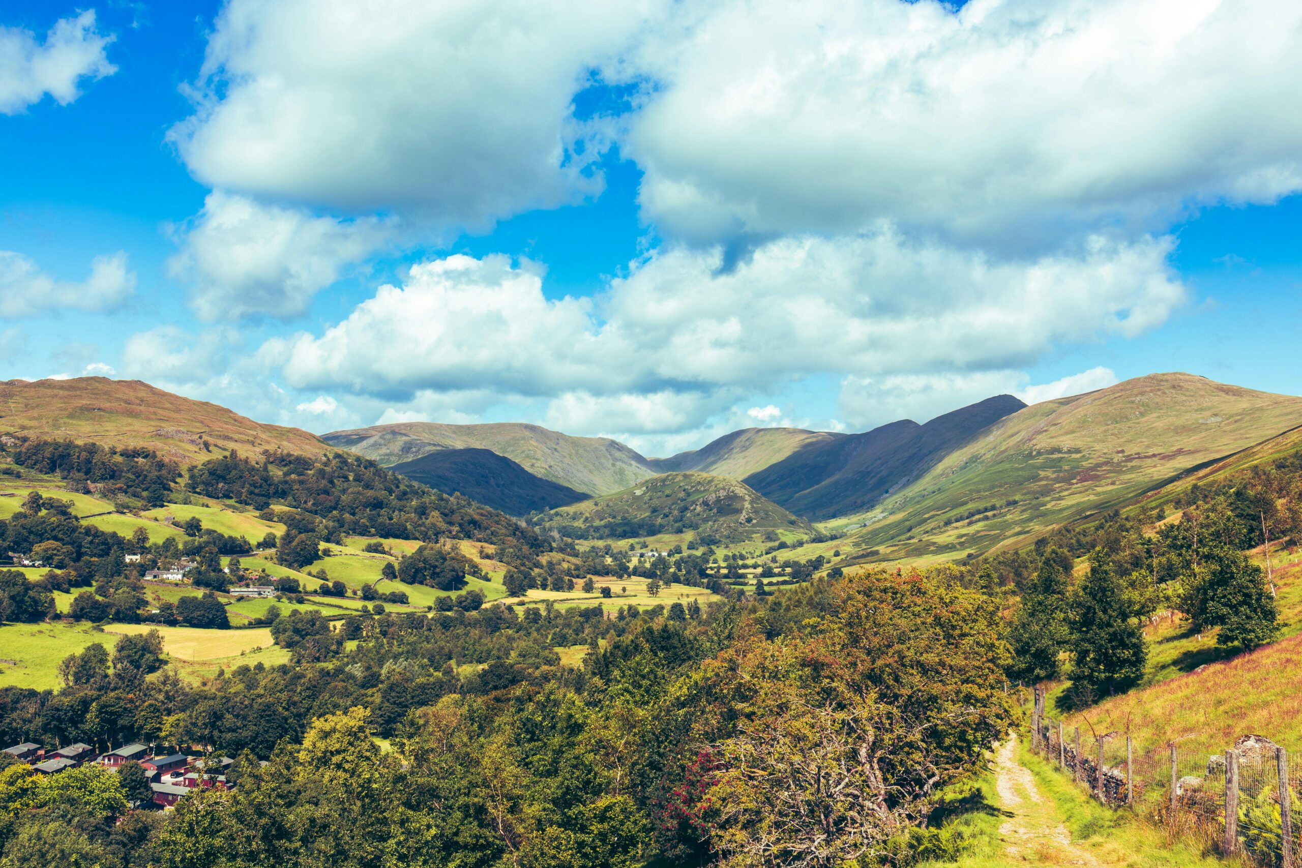 A sweeping view across a lush green Lake District valley, with patchwork fields, scattered farmhouses and woodland in the foreground giving way to dramatic fells rising under a bright blue sky dotted with large white clouds