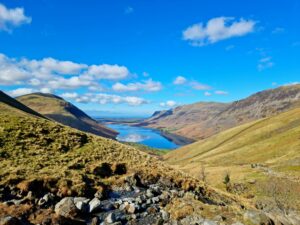 A sweeping valley view looking down Wasdale valley in the Lake District. Wast Water is nestled between rolling hills and rugged mountains under a bright blue sky with scattered white clouds. In the foreground, a rocky stream bed cuts through golden-brown moorland grass.