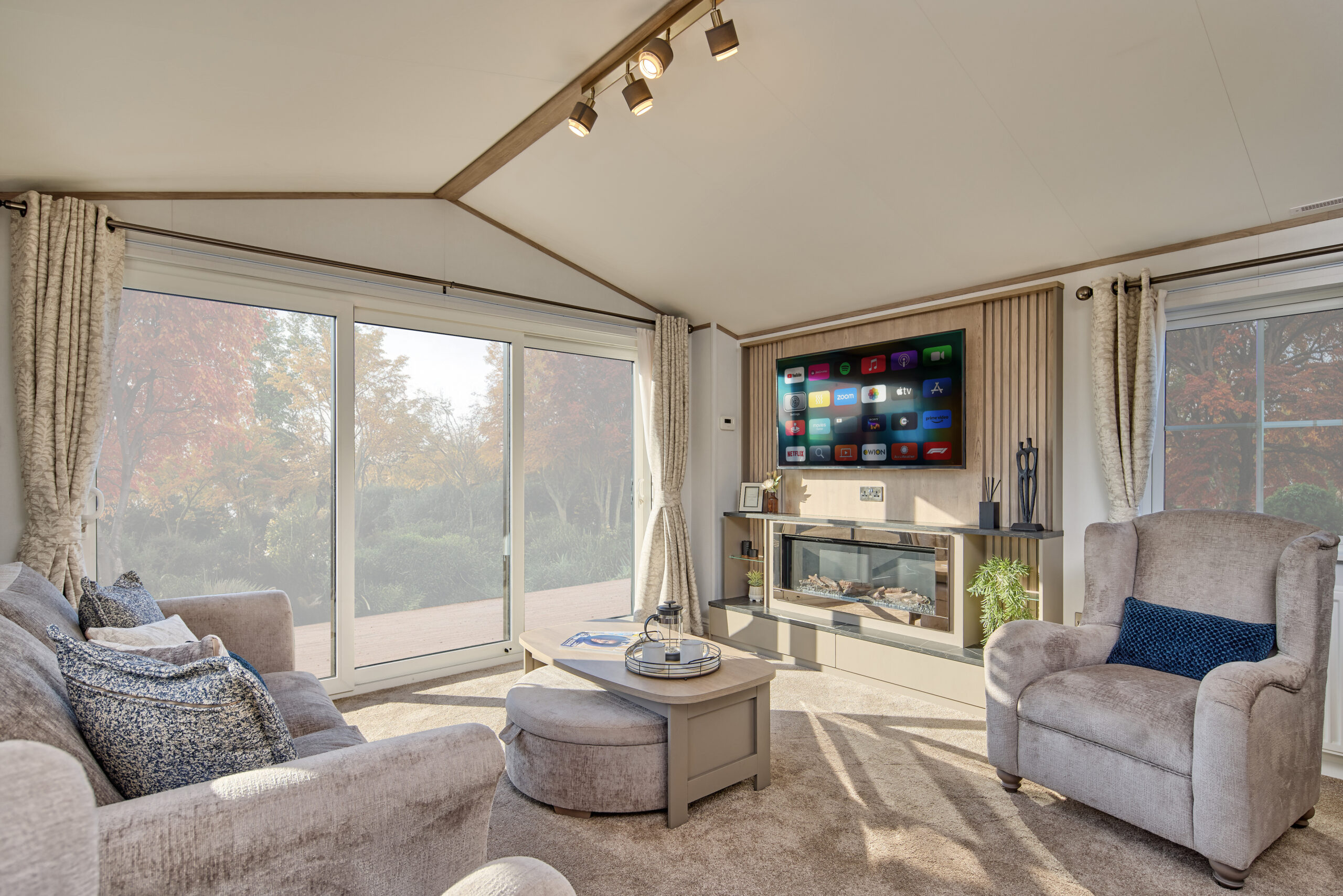 Spacious living room in the Willerby Vogue static caravan, featuring plush sofas, a wall-mounted smart TV above a fireplace, and full-length windows looking out onto autumnal woodland.