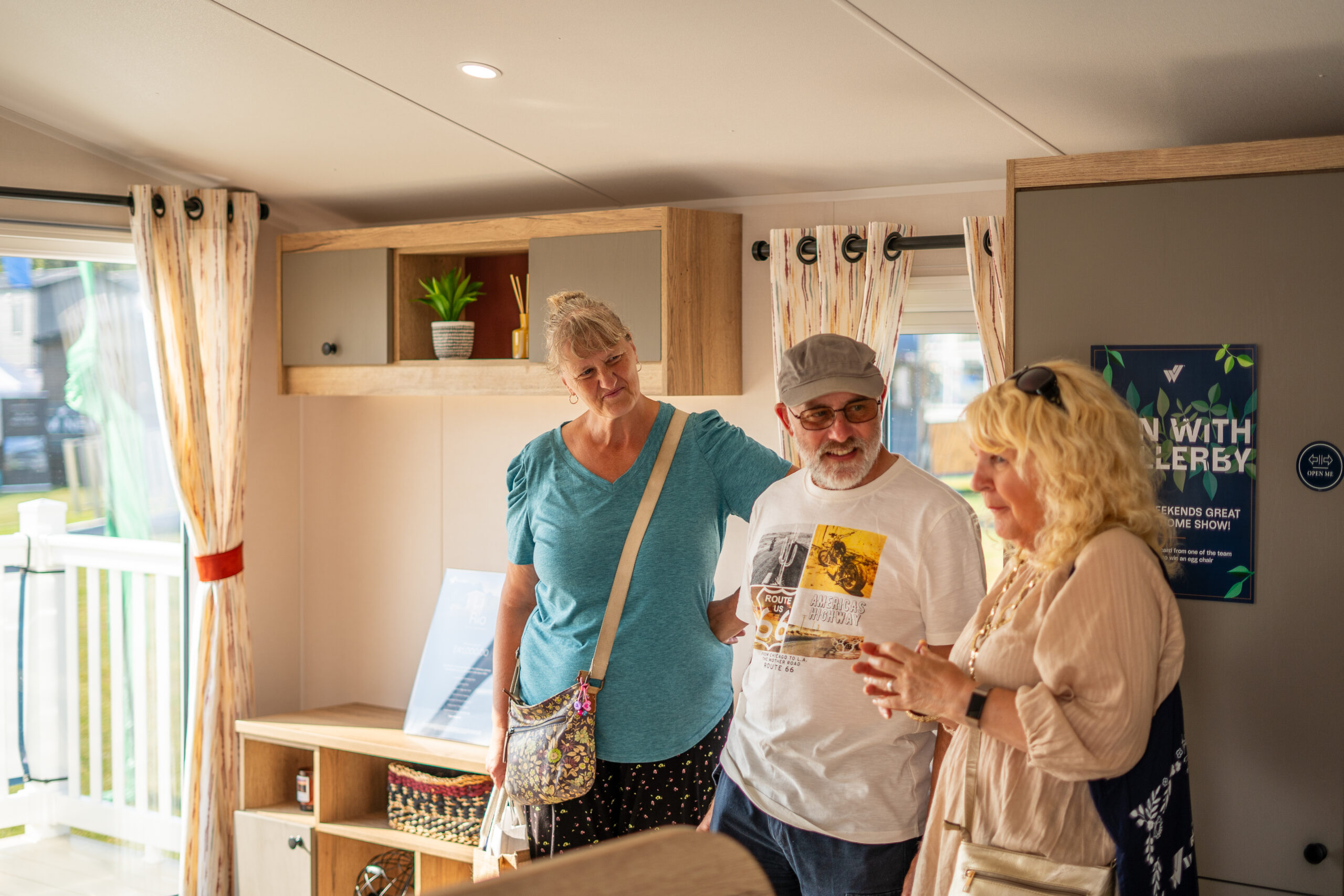 Visitors chatting inside a Willerby holiday home show home, with wooden storage units, patterned curtains, and a promotional poster visible in the background.