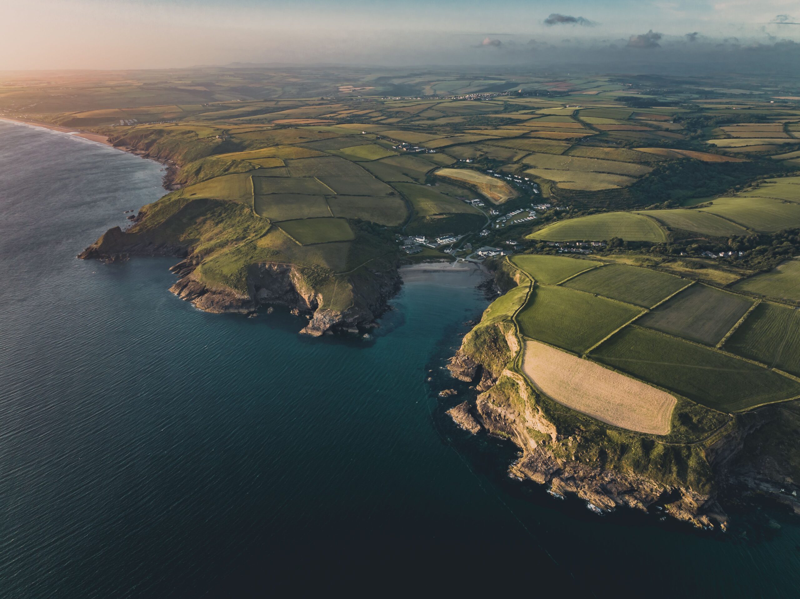 An aerial photograph of the Pembrokeshire coastline capturing a breathtaking view of the ocean meeting lush green farmland. The clear blue ocean contrasts with the rolling green hills of the countryside, which are divided into patchwork squares.