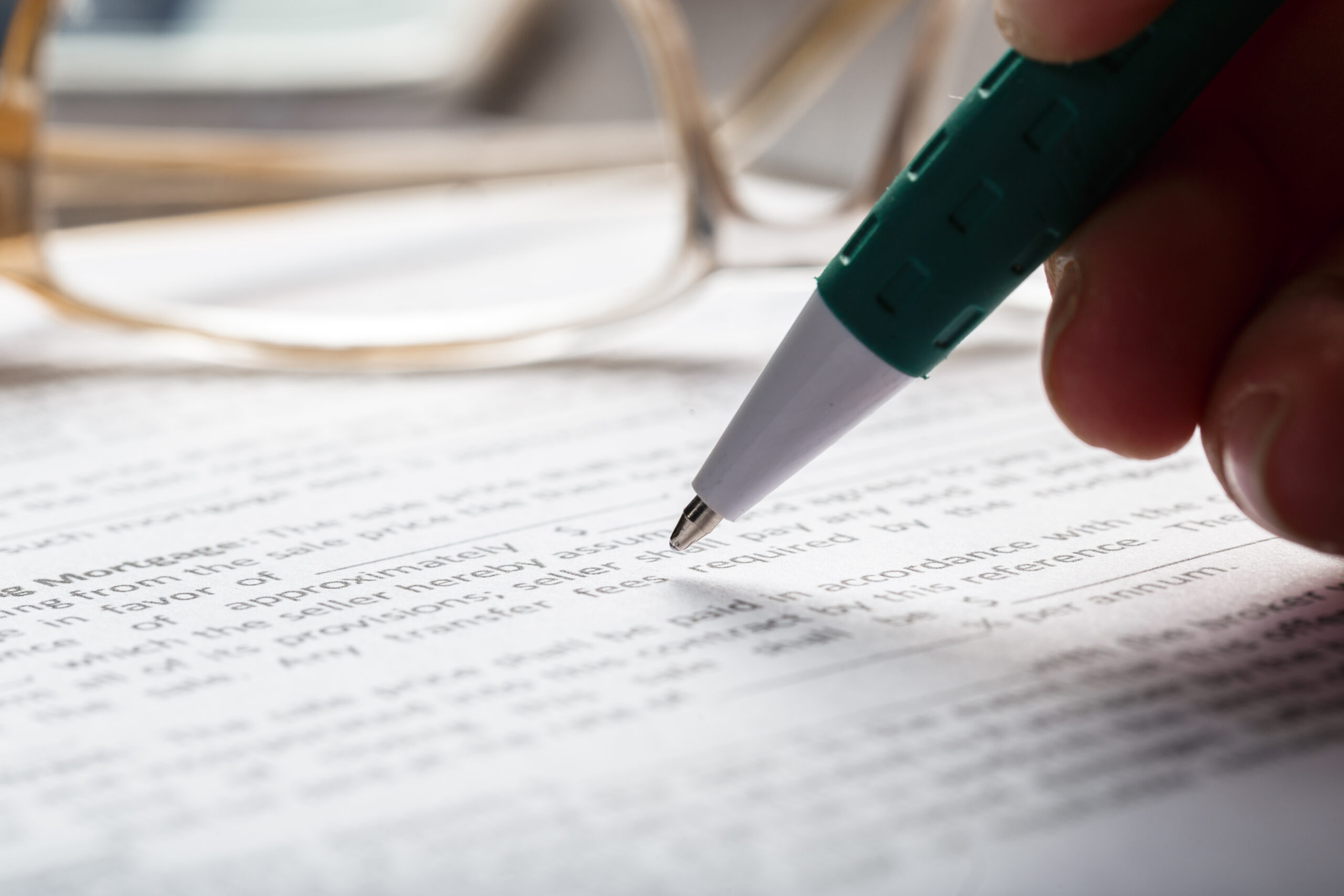 Close-up of a hand holding a green and white pen, poised over a printed mortgage document, with a pair of eyeglasses resting on the desk in the softly blurred background.