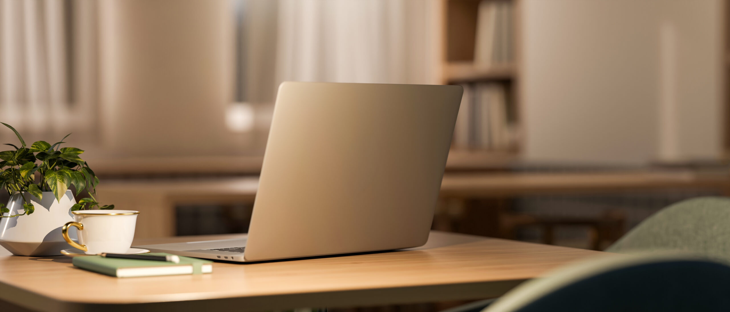 An open laptop sits on a wooden desk, viewed from behind, with a gold-rimmed white teacup, a small potted plant, a green notebook, and a pen nearby. Blurred bookshelves and warm, soft-focus curtains fill the background.