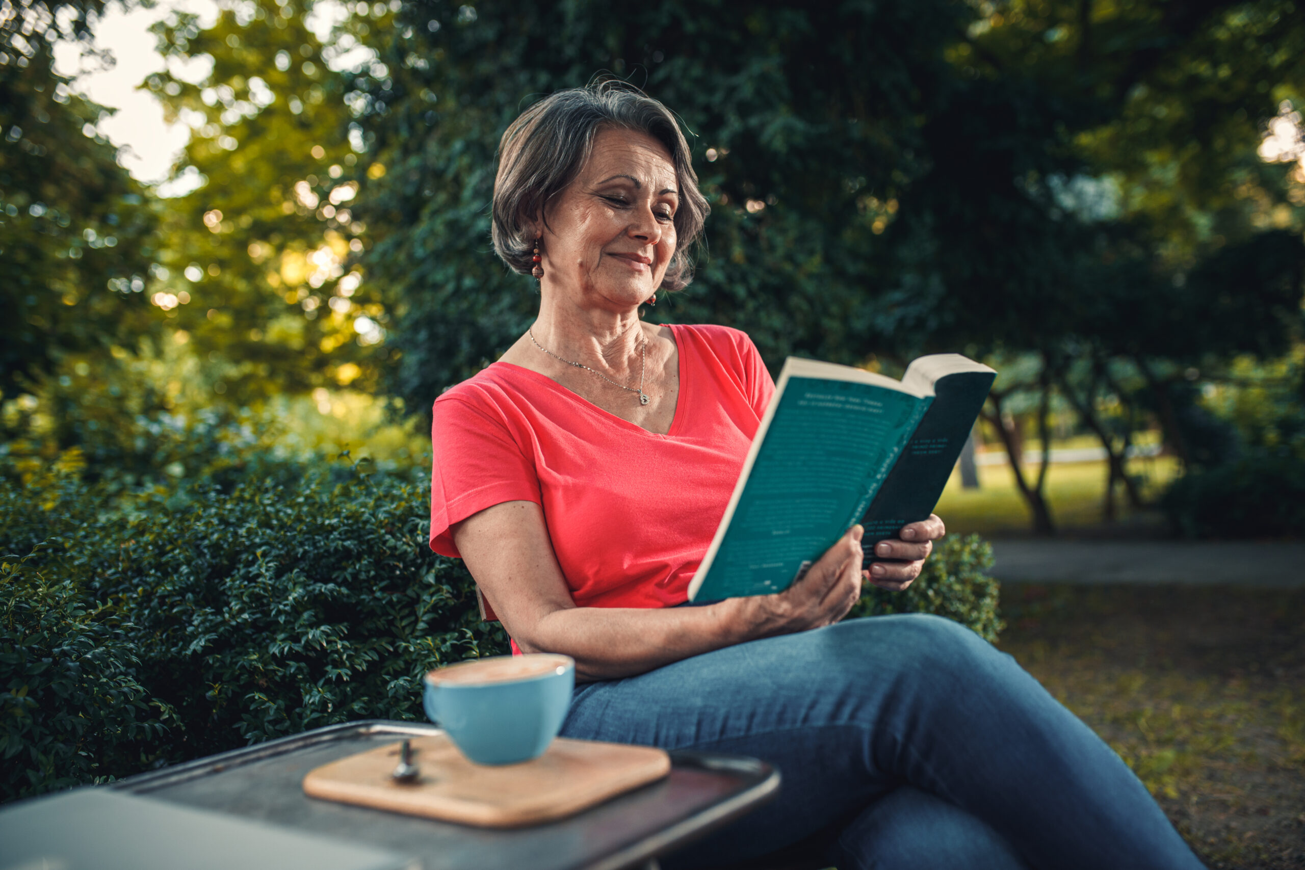 A senior woman with short grey hair sits outdoors, reading a book and enjoying a cup of coffee. She is wearing a red shirt and jeans, and a necklace. The table next to her holds a blue mug and a wooden tray. The background shows greenery.
